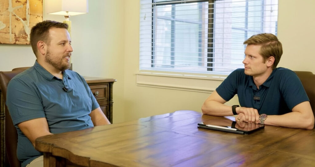 Two men, wearing blue shirts, sitting across a wooden table from each other in an office setting. They are engaged in a professional discussion or interview.